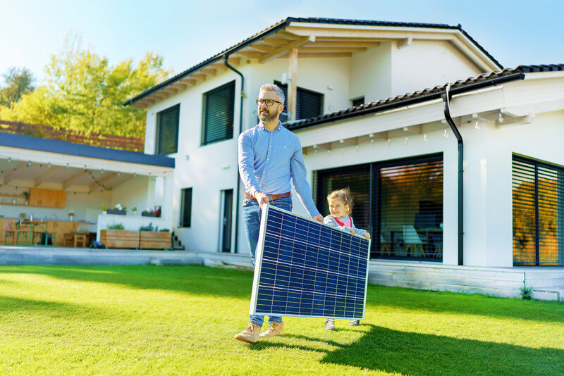 Father With His Little Daughter Carring Solar Panel At Their Backyard. Alternative Energy, Saving Resources And Sustainable Lifestyle Concept. Father With His Little Daughter Carring Solar Panel At Their Backyard. Alternative Energy, Saving Resources And Sustainable Lifestyle Concept.